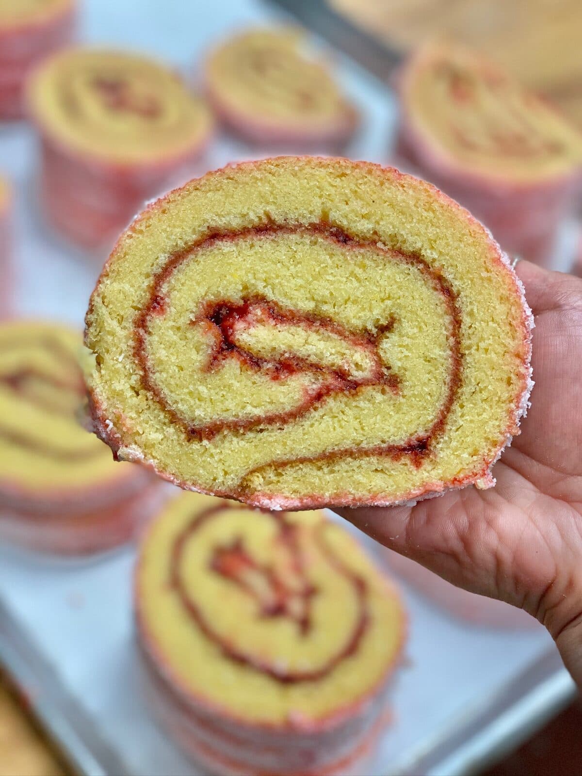 Cross-section of a niño envuelto roll cake with strawberry swirl, held above a tray of more rolls
