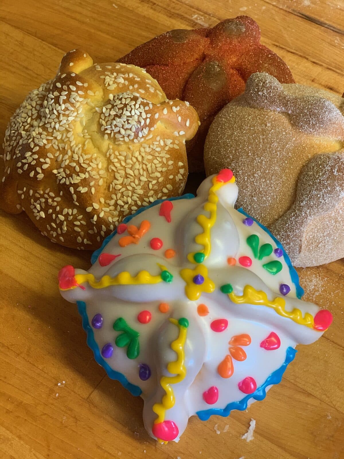 Four styles of pan de muerto: sesame-seed, sugar-coated, plain, and colorfully iced, arranged on a wooden bakery table
